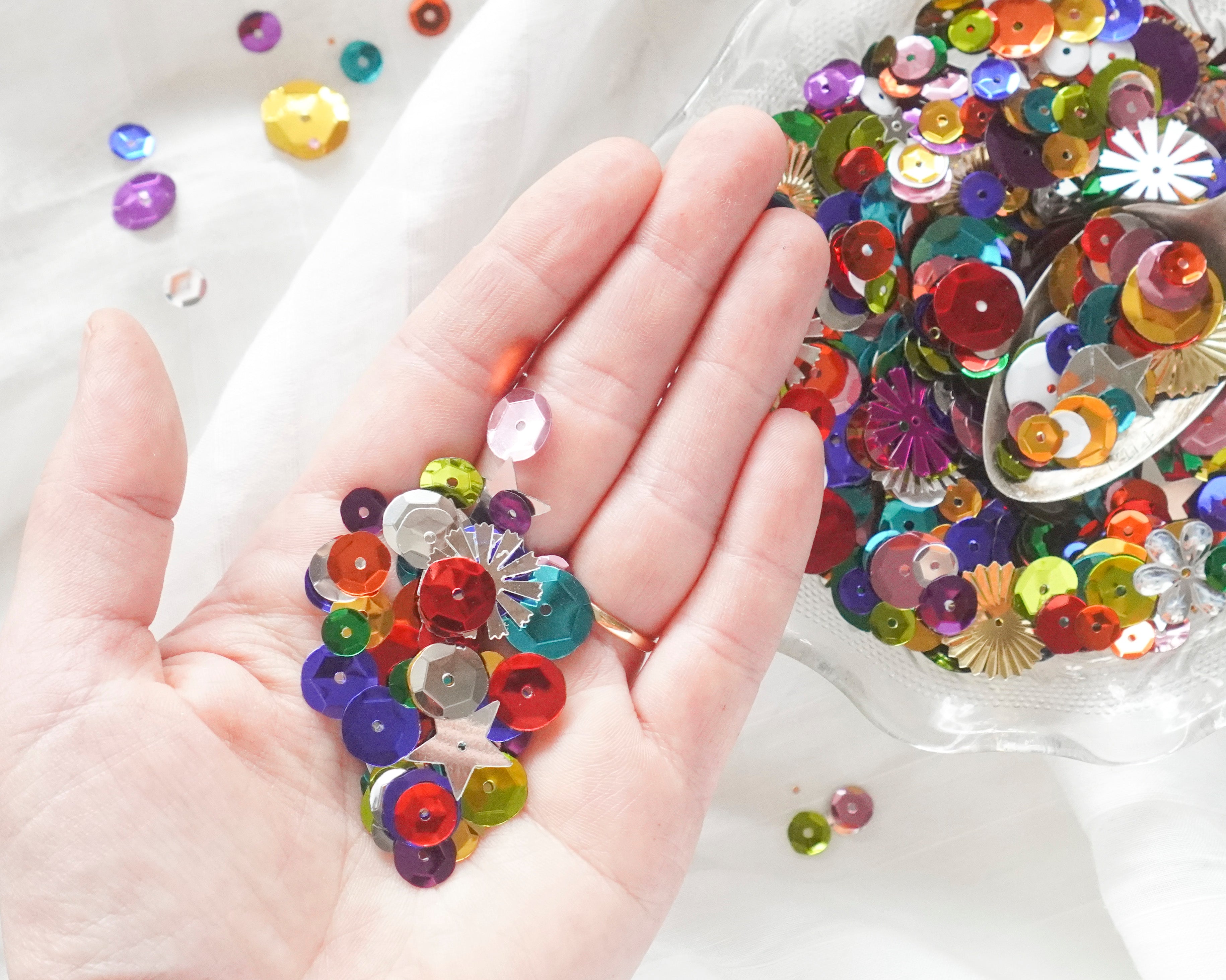 Colorful sequins on a hand with a clear plastic bowl of sequins in the background.