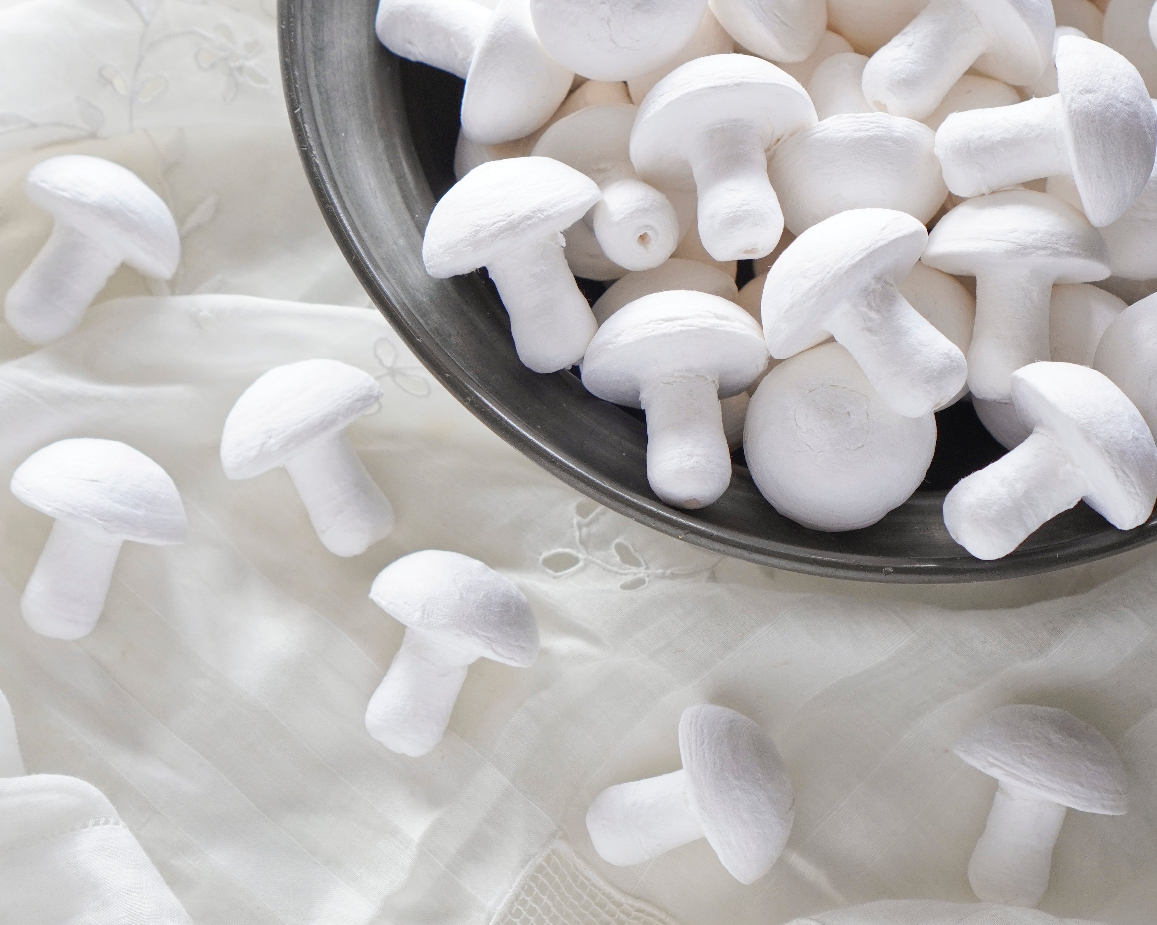 White spun cotton craft mushrooms arranged on a light fabric surface, with several clustered inside a dark metal bowl.