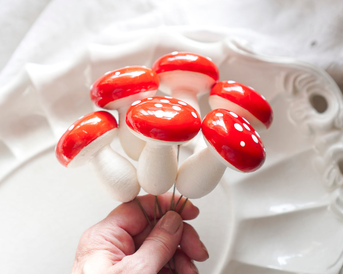 A hand holding a collection of red-and-white mushroom decorations with glossy red caps and white stems, photographed against a soft white ceramic and fabric background.