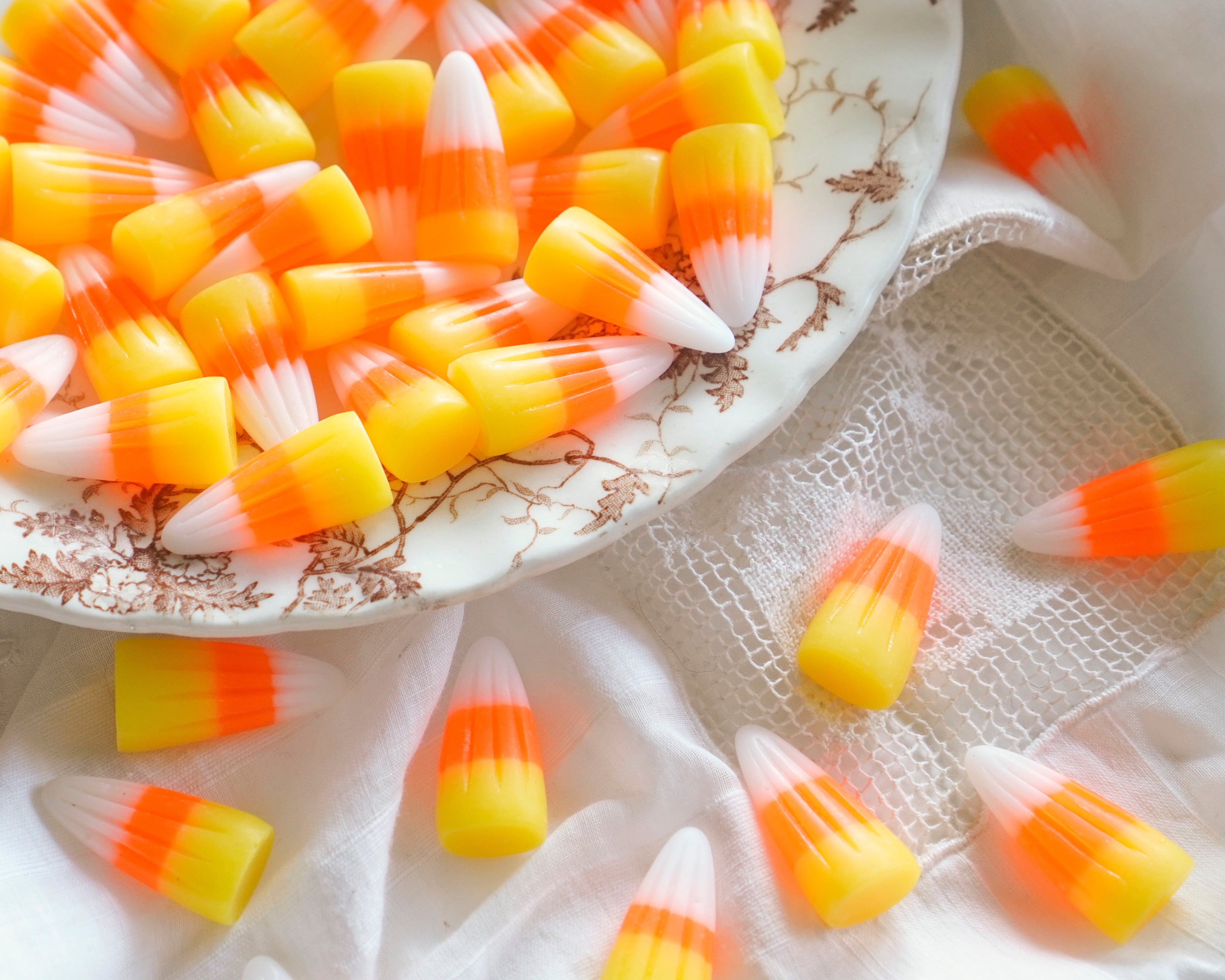 Candy corn pieces arranged on a white plate with a floral rim and scattered on white fabric, showing the classic yellow, orange, and white color bands.