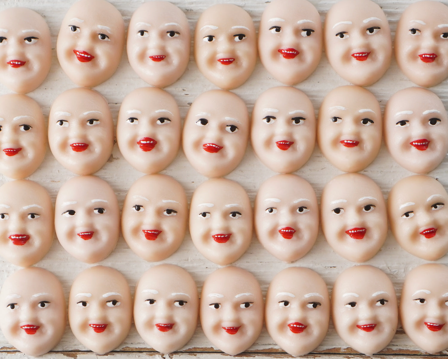 An array of miniature plastic face cabochons in a cream color, arranged in rows, with each face featuring painted details such as eyes, mouth, and red accents.