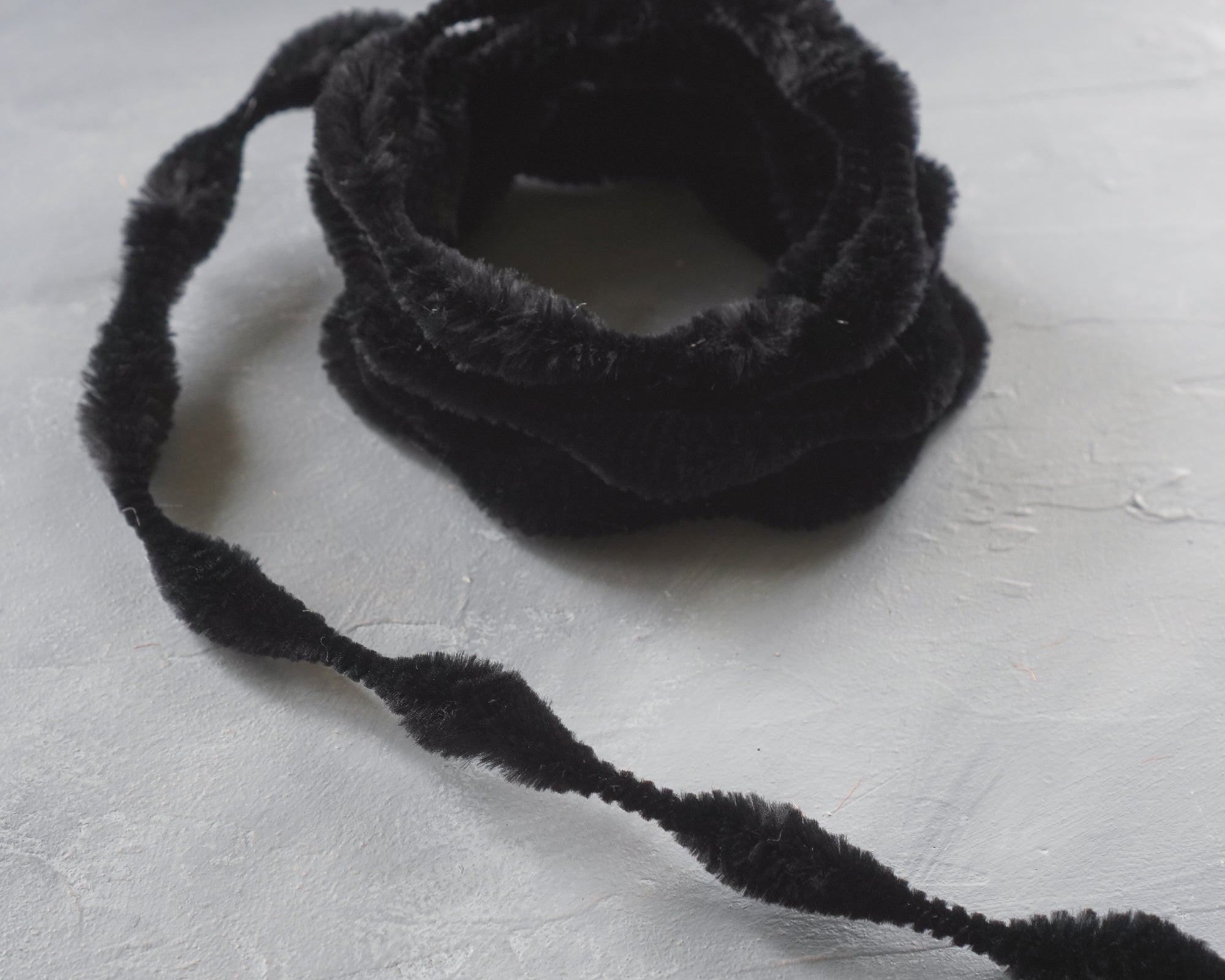 A coil of black bump chenille trim on a white surface, with the trim uncoiled to show the texture and thickness.