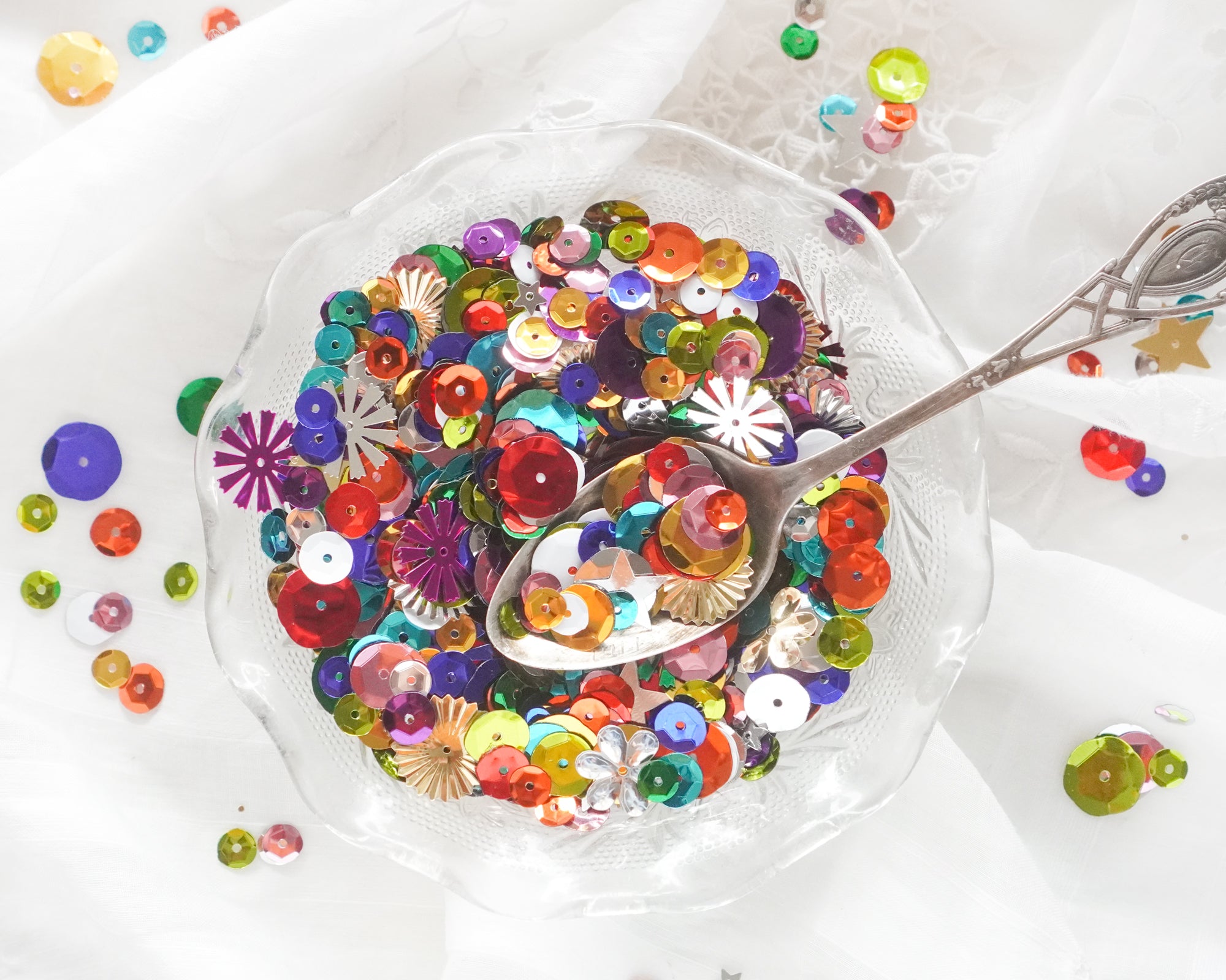 Colorful sequins in a clear bowl with a spoon on a white background