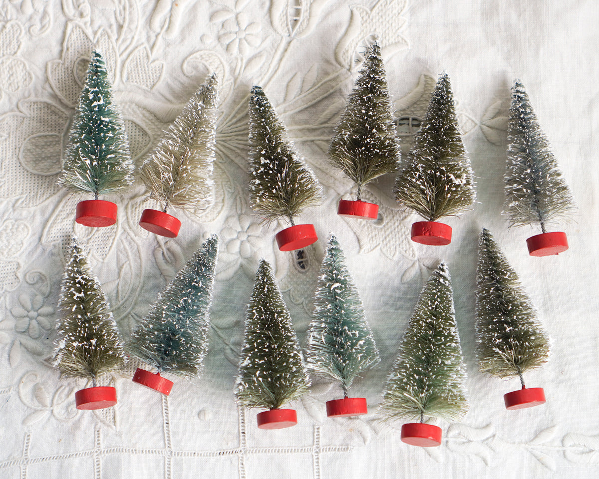Miniature bottle brush trees with red bases displayed on a white cloth.