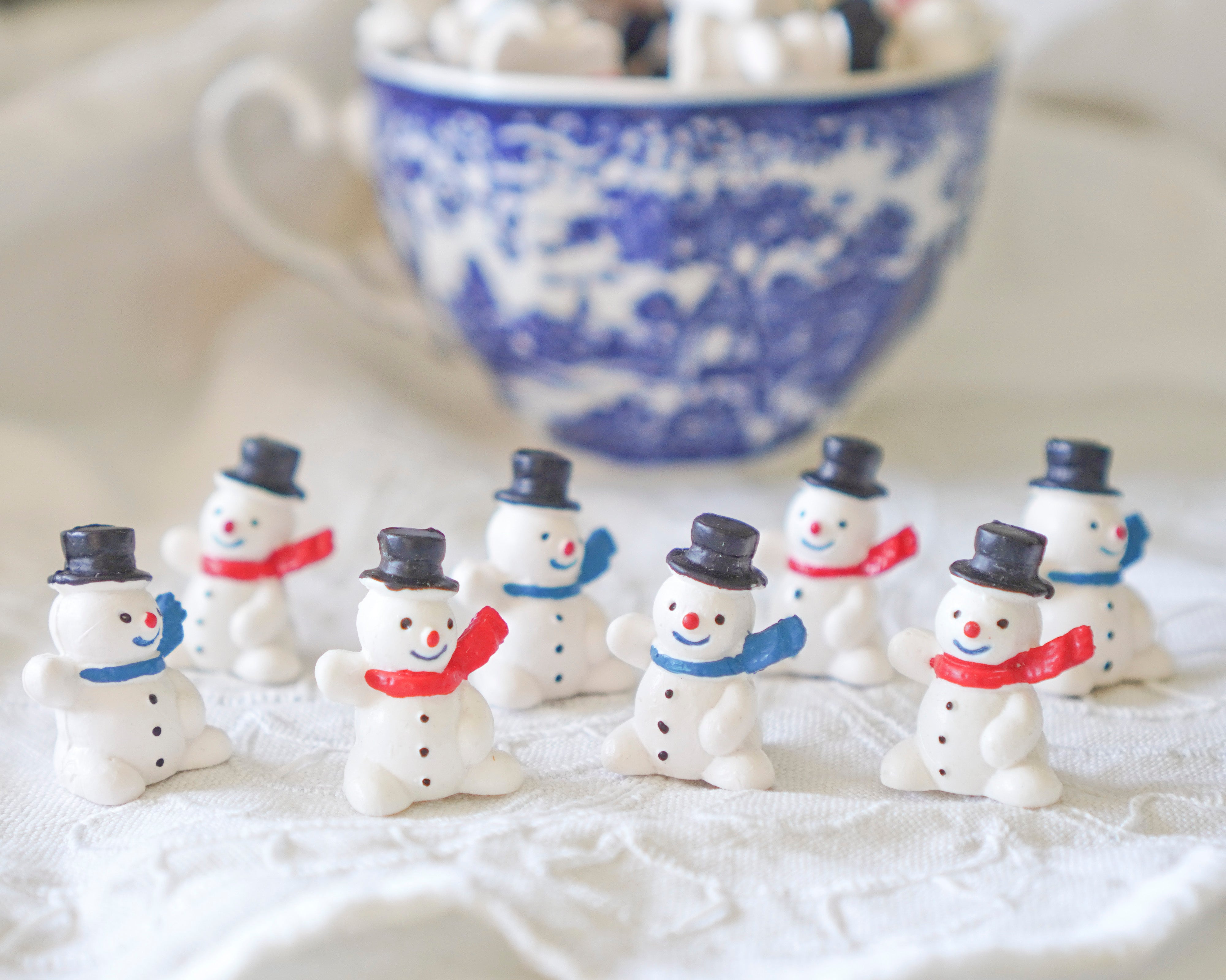 Small snowman figurines with top hats and scarves on a white surface with a blue and white patterned cup in the background.