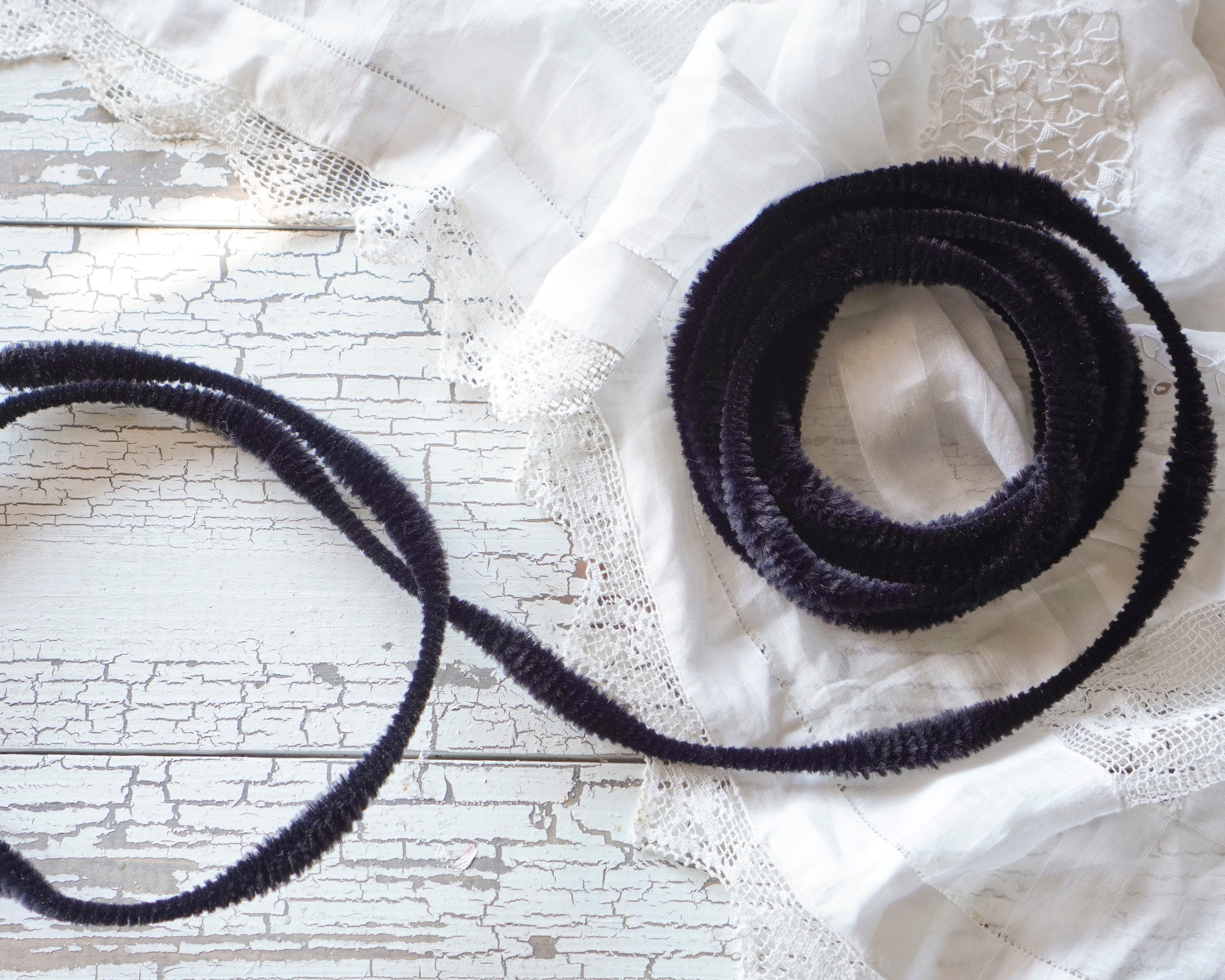 A coiled black fuzzy bump chenille trim ribbon laid on a white distressed wood surface with white lace fabric draped around it.