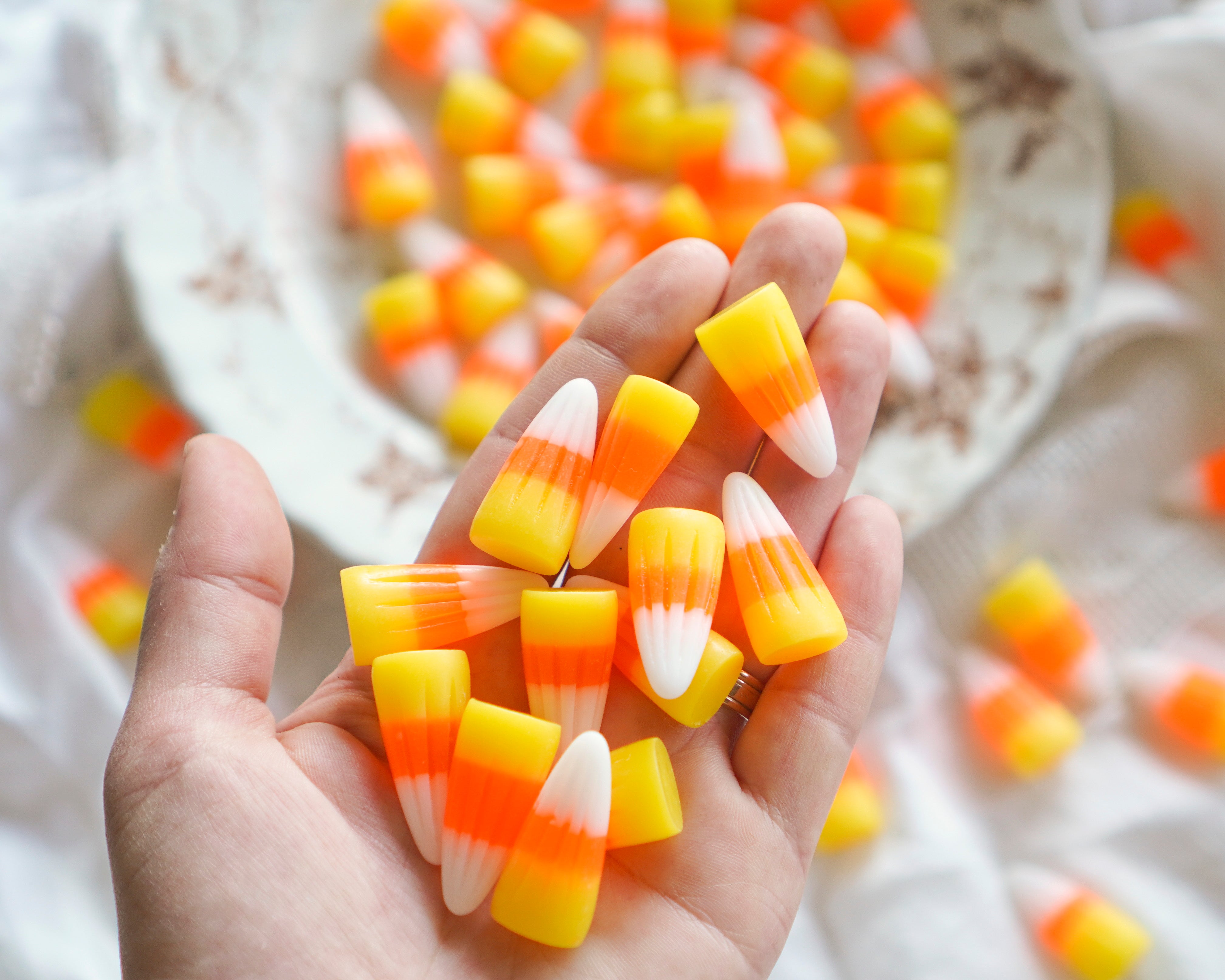 A hand holding several candy corn decorations, with more candy corn pieces scattered on a white fabric background and a blurred bowl in the back.