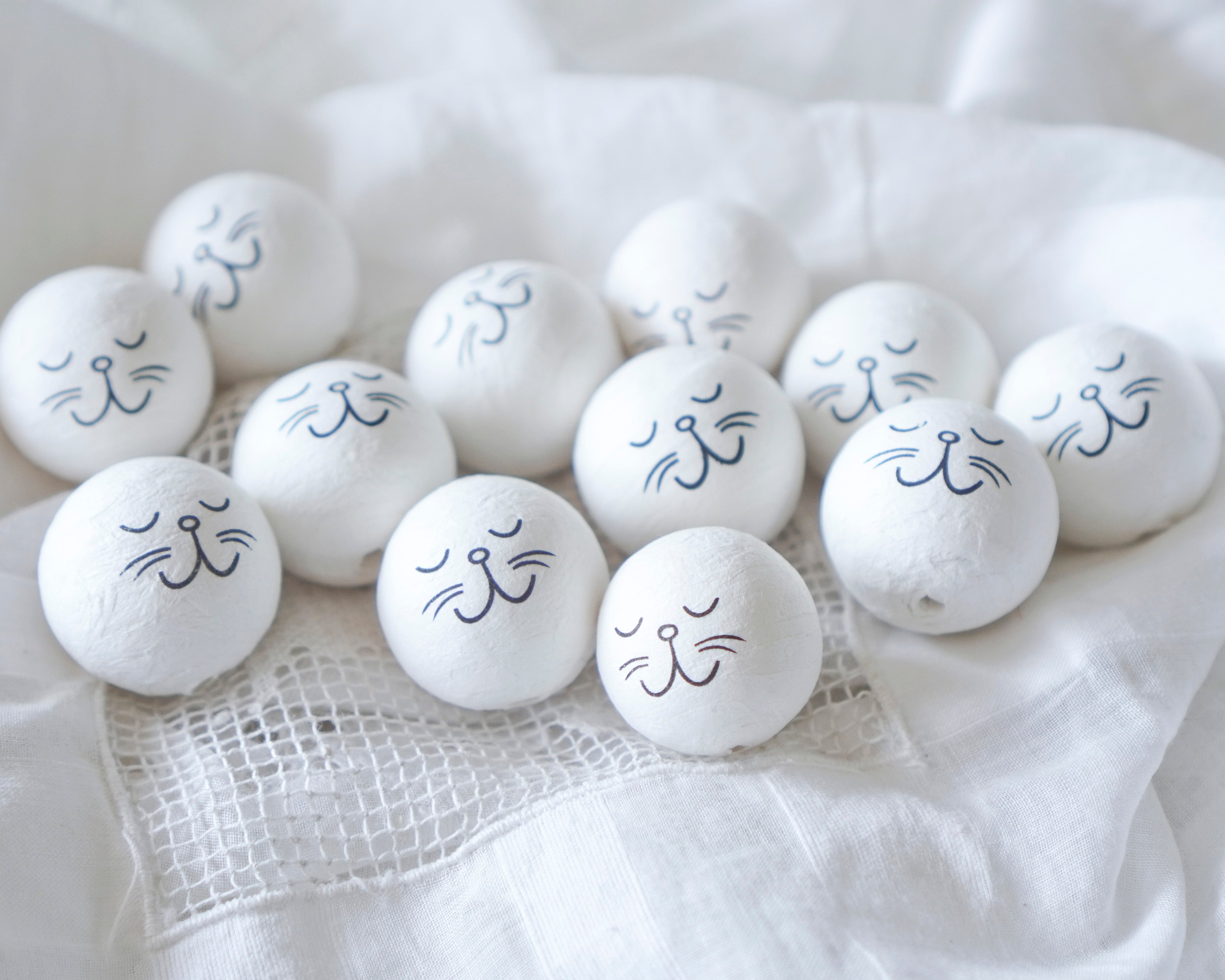 A group of white bunny craft heads decorated with simple black cat face drawings, arranged on soft white fabric.