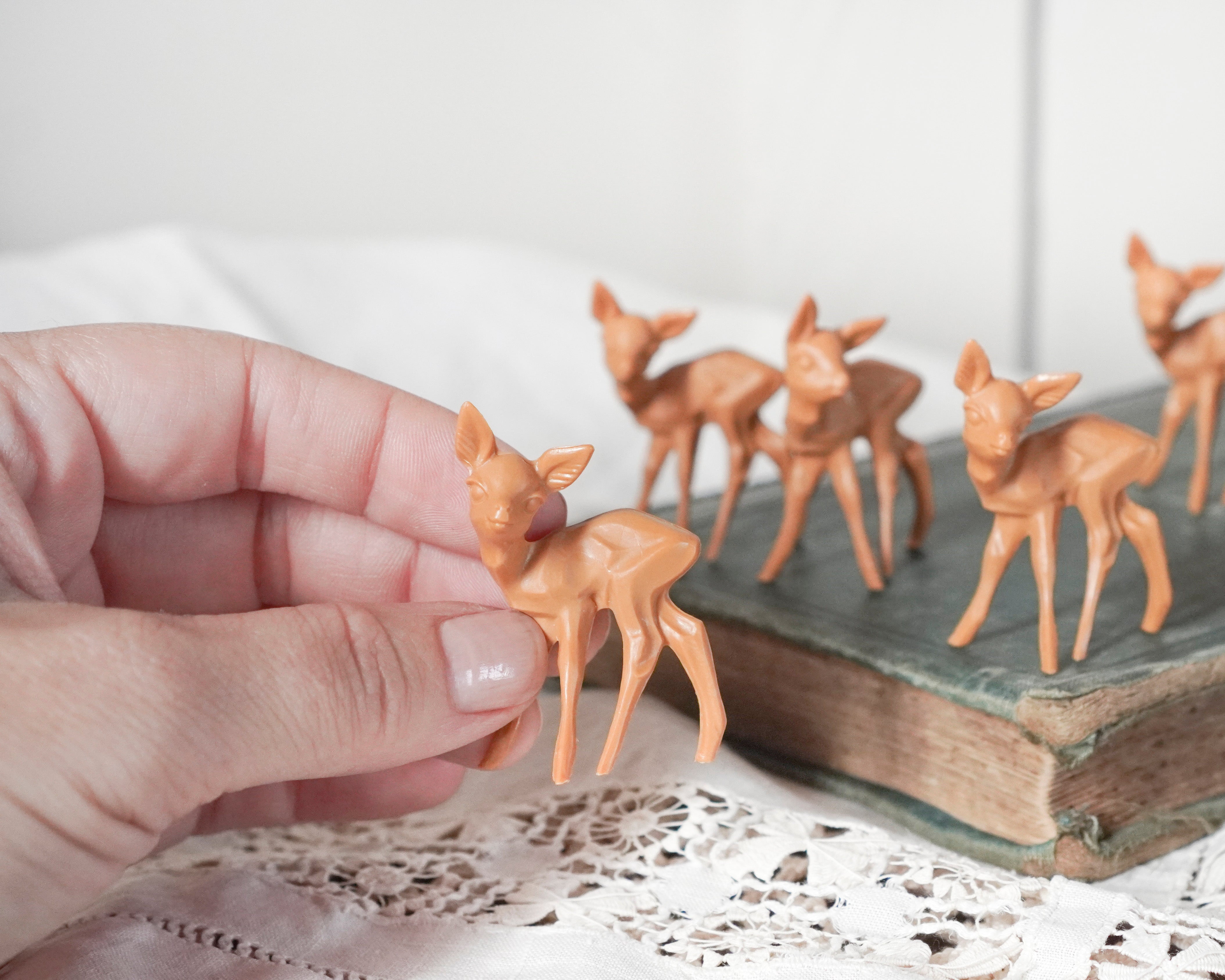 A hand holds a small tan deer figurine in the foreground, with several matching deer figurines displayed on an old book in the background.