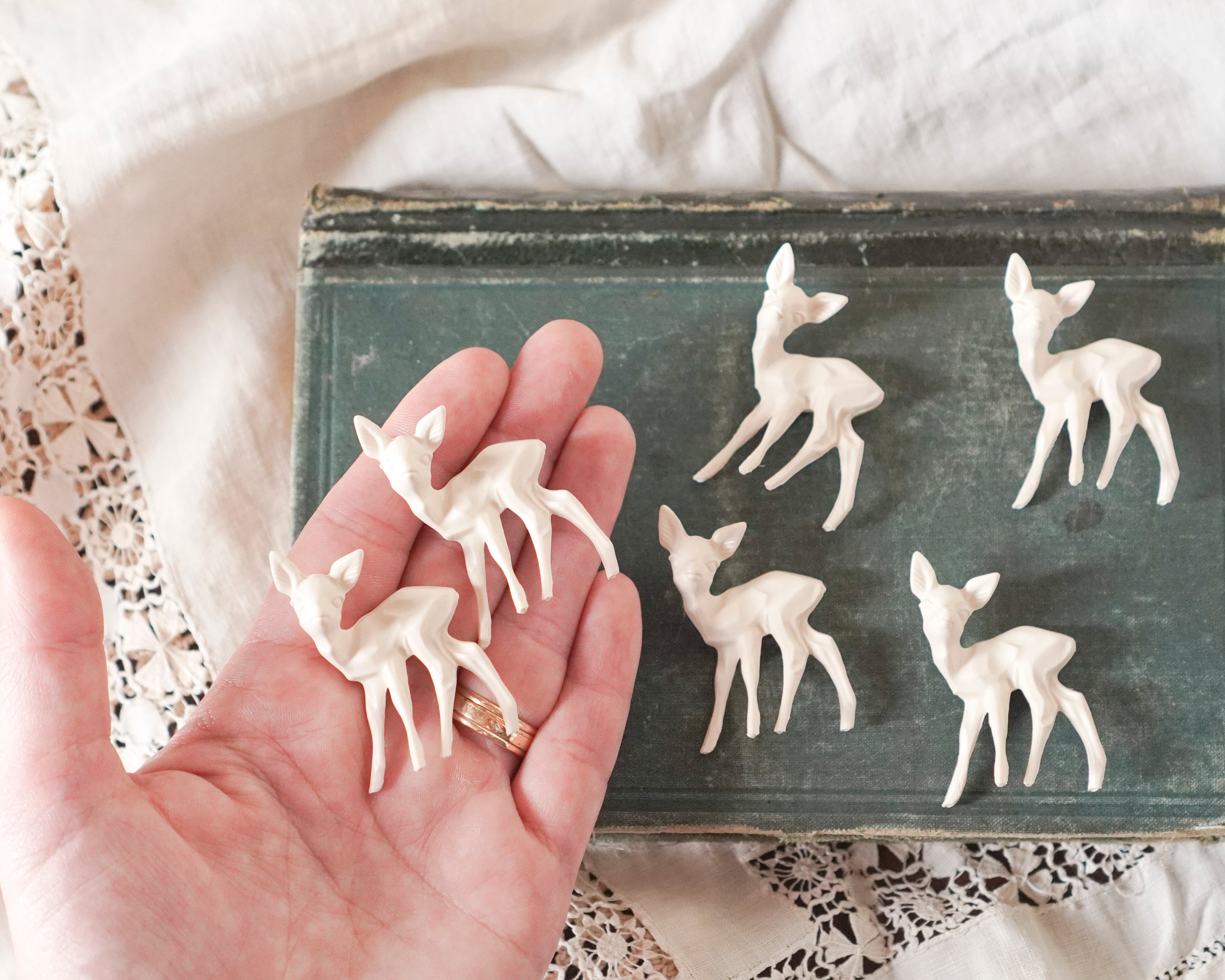 A hand holds small cream deer figurines, with more placed on a distressed dark green book. The scene is styled with cream fabric and lace.