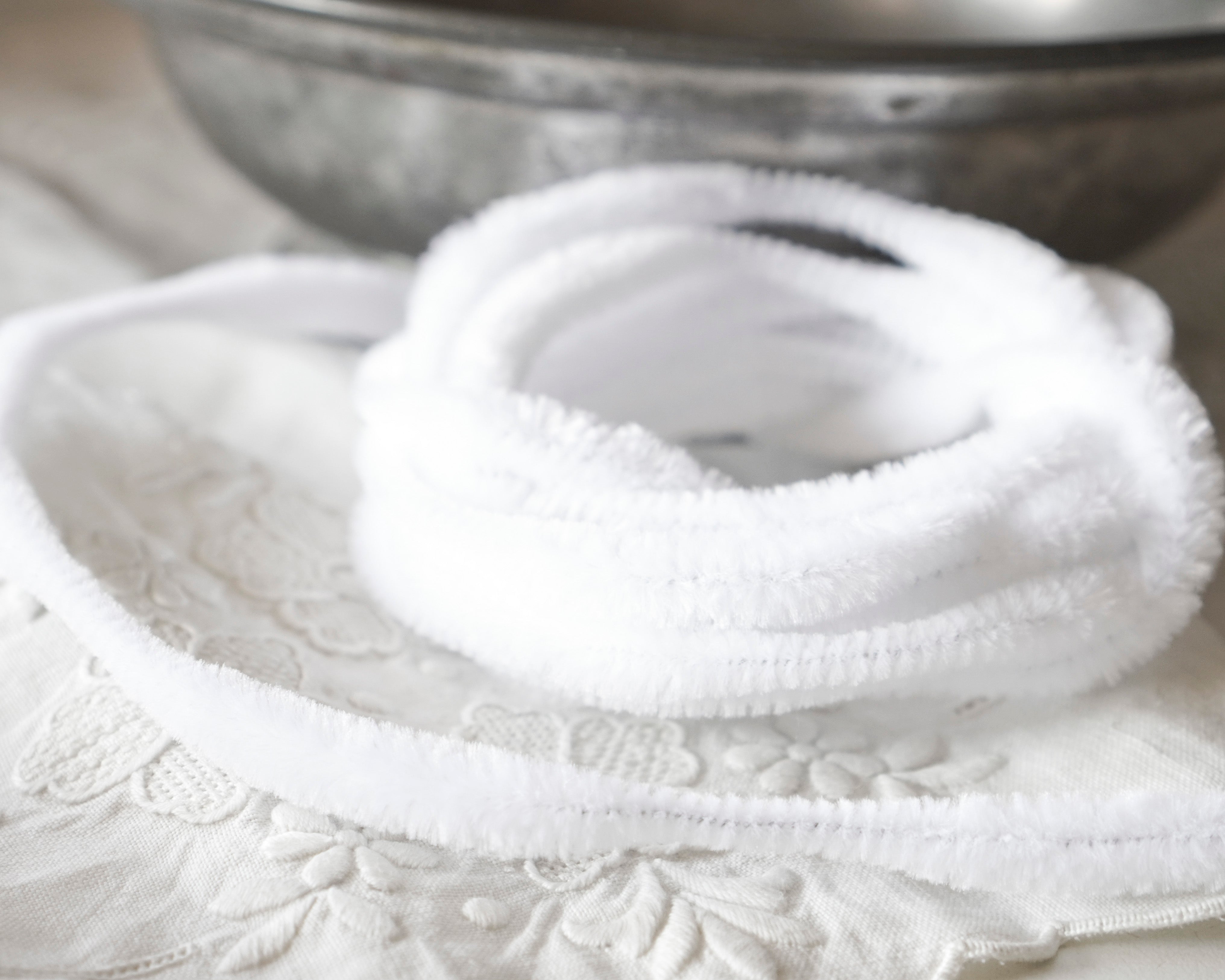 White fuzzy bump chenille craft trim coiled on a white embroidered cloth, with a blurred metal bowl in the background.